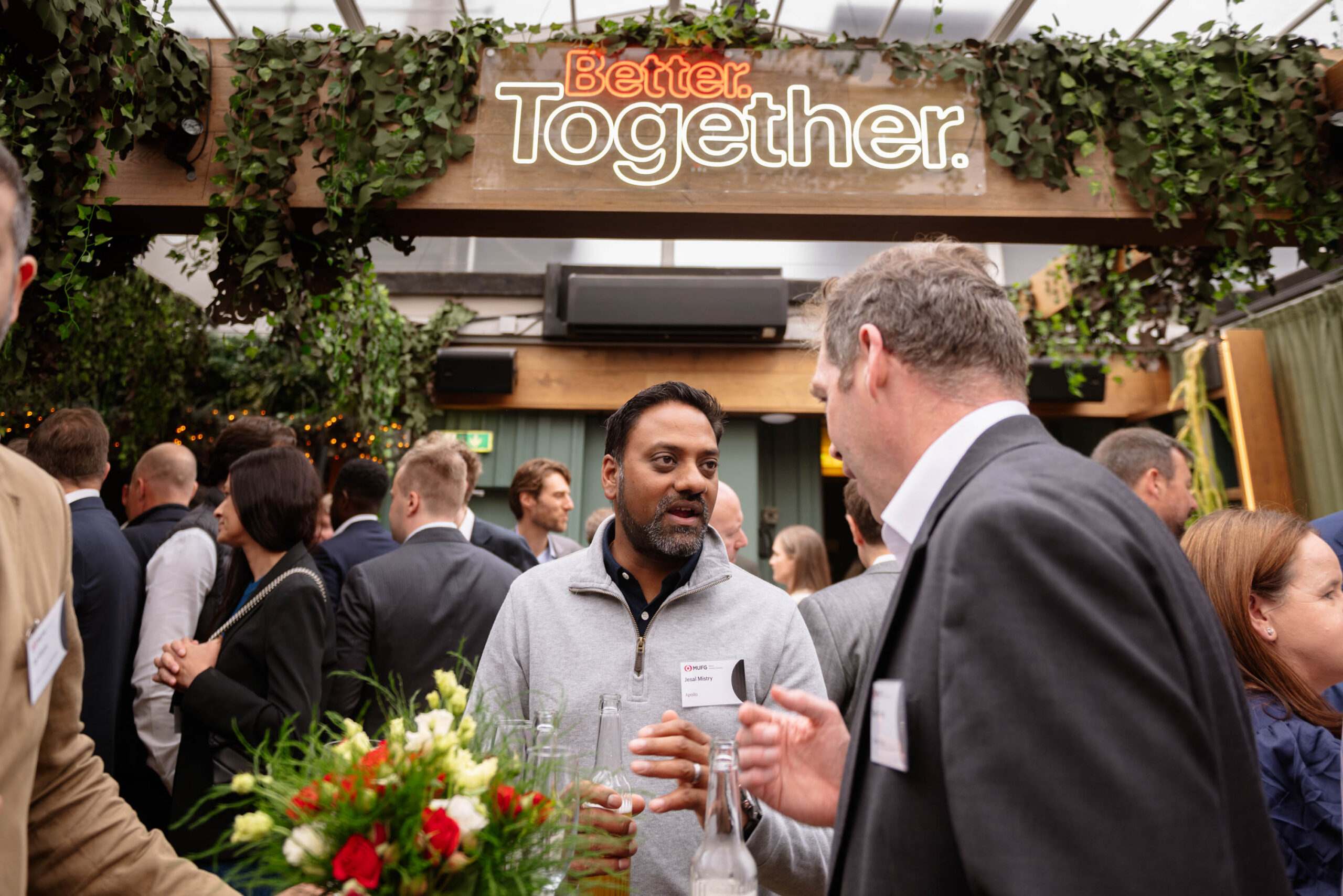 Group of well-dressed people at a social event around a floral centerpiece, under a neon 'Better Together' sign.