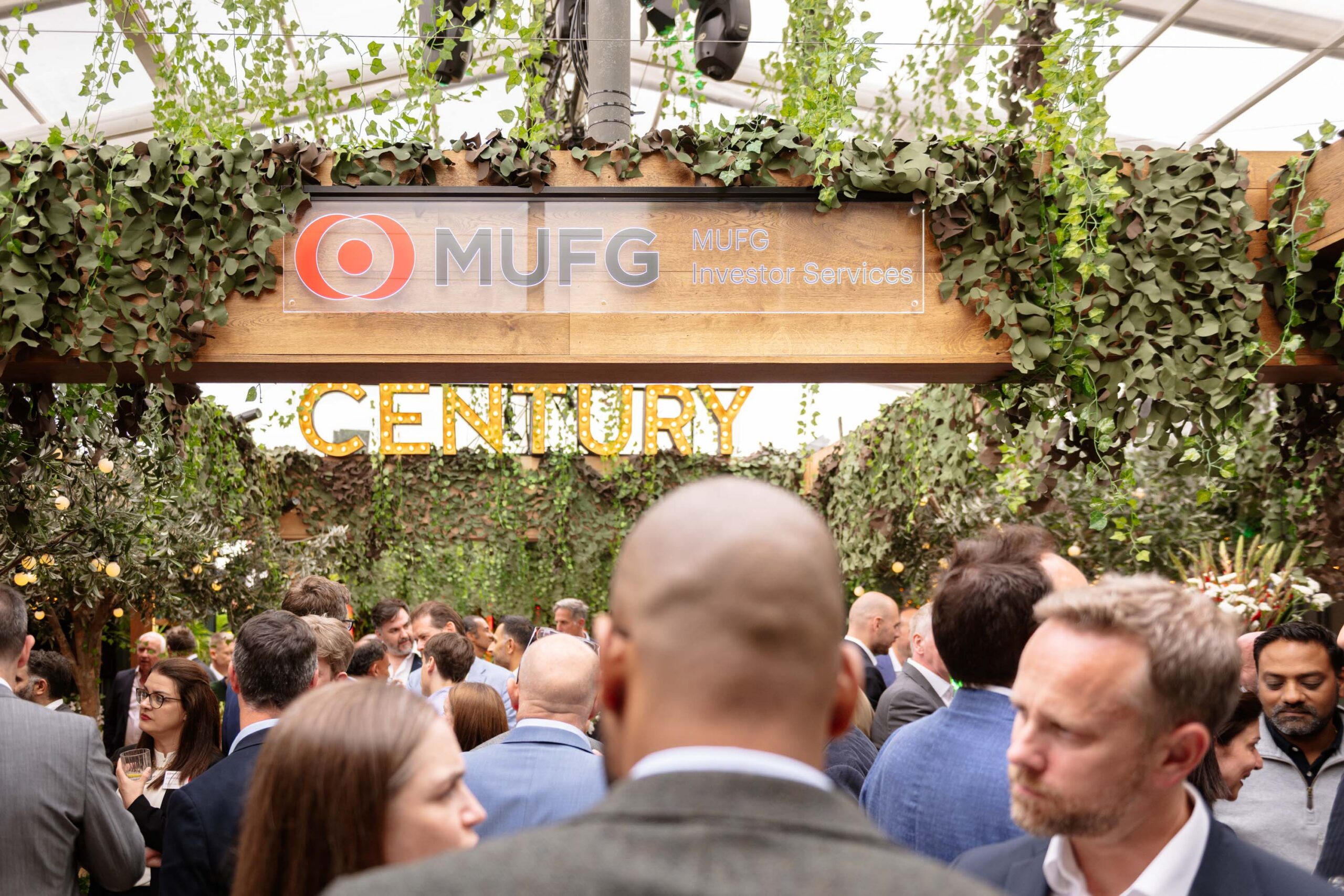 Crowded indoor event under a leafy canopy with a wooden sign reading MUFG Investor Services.