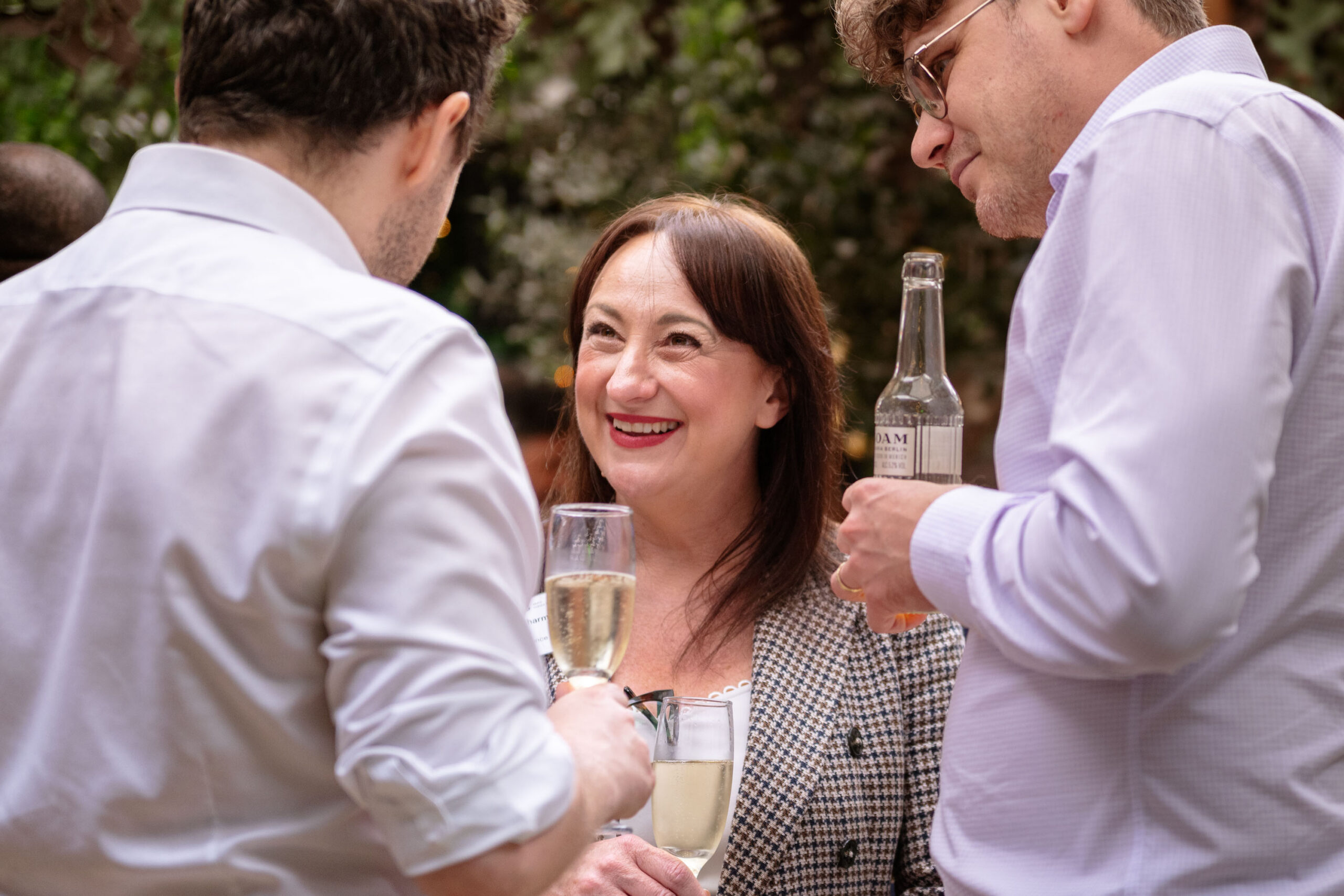 Woman in a checkered blazer smiles at two men in white shirts outdoors, holding a flute of champagne amid a social gathering.
