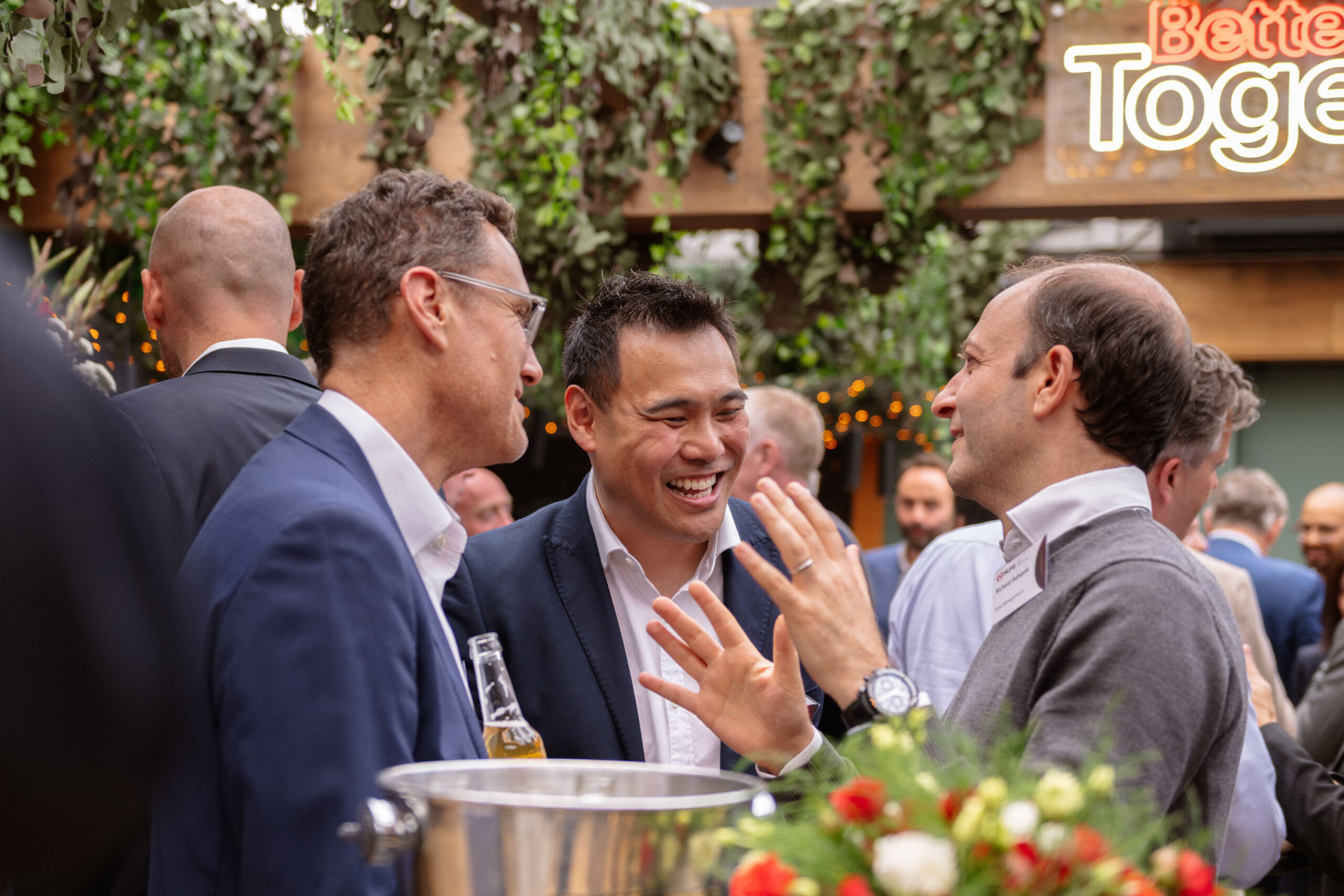 Group of professionally dressed men socializing at a reception with drinks and greenery in the background.