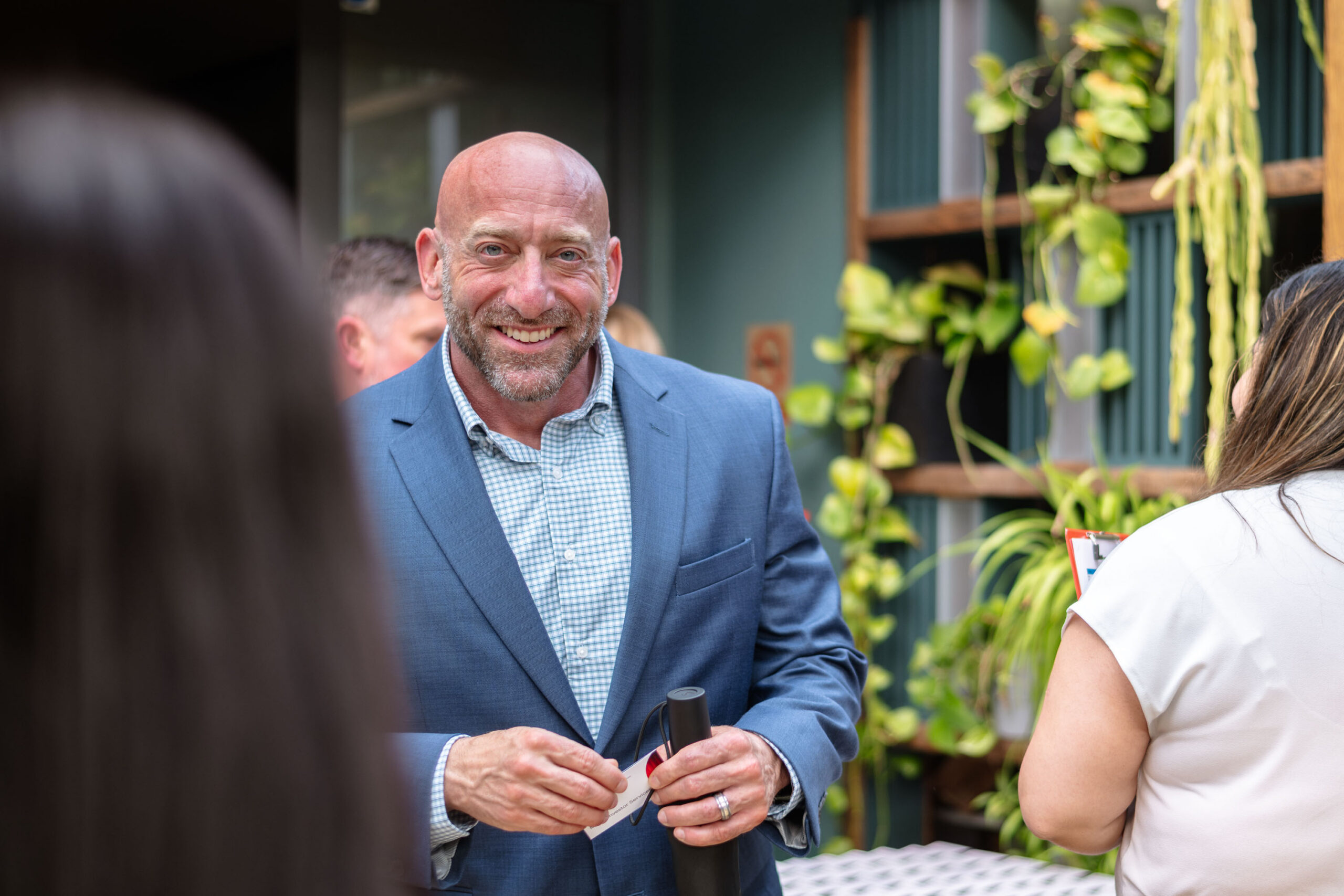 Smiling bald man in a blue blazer talks with someone at an outdoor event, holding papers and glasses.