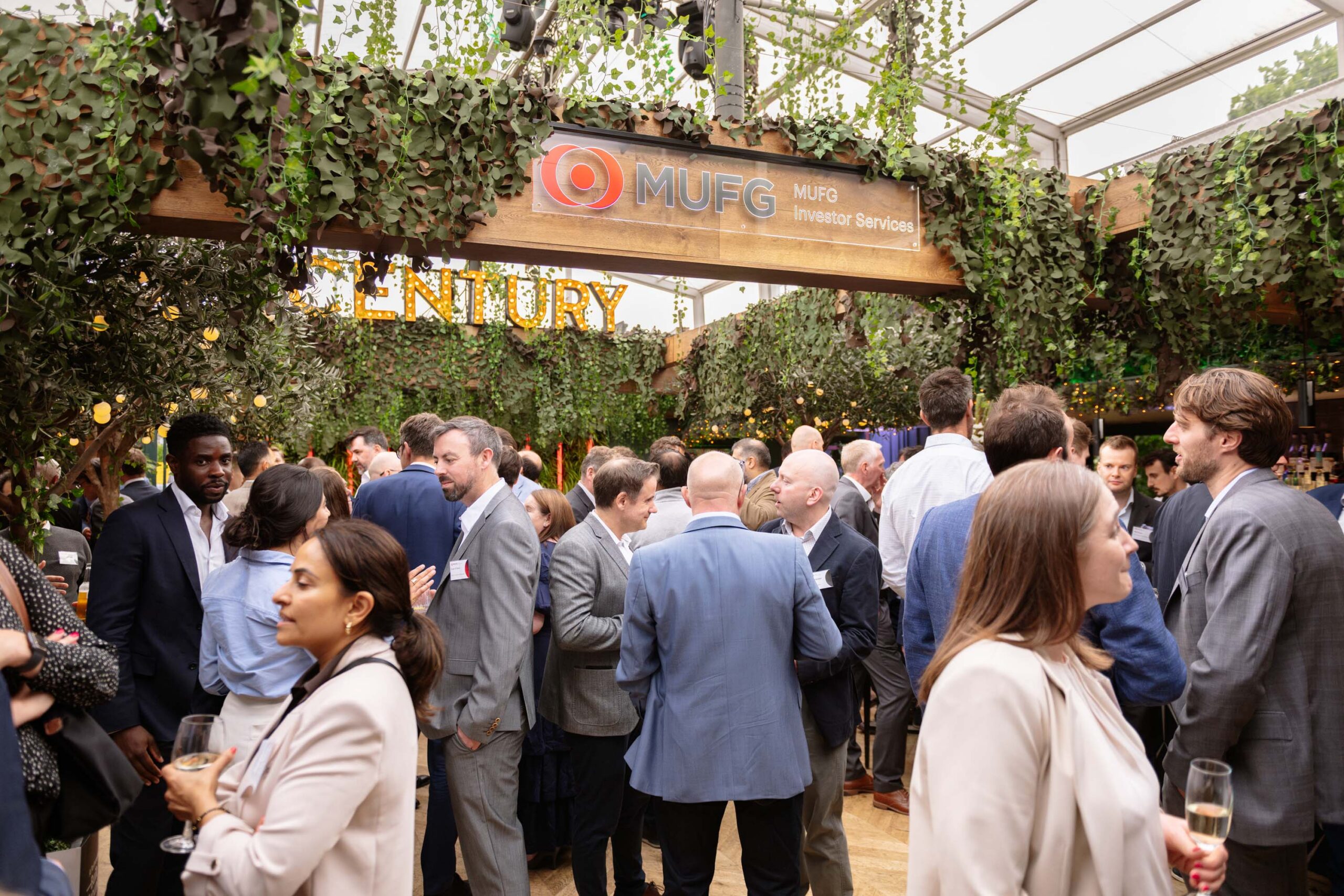 Professionals in suits networking at a leafy corporate event under ivy-covered beams with a MUFG Investor Services sign above.
