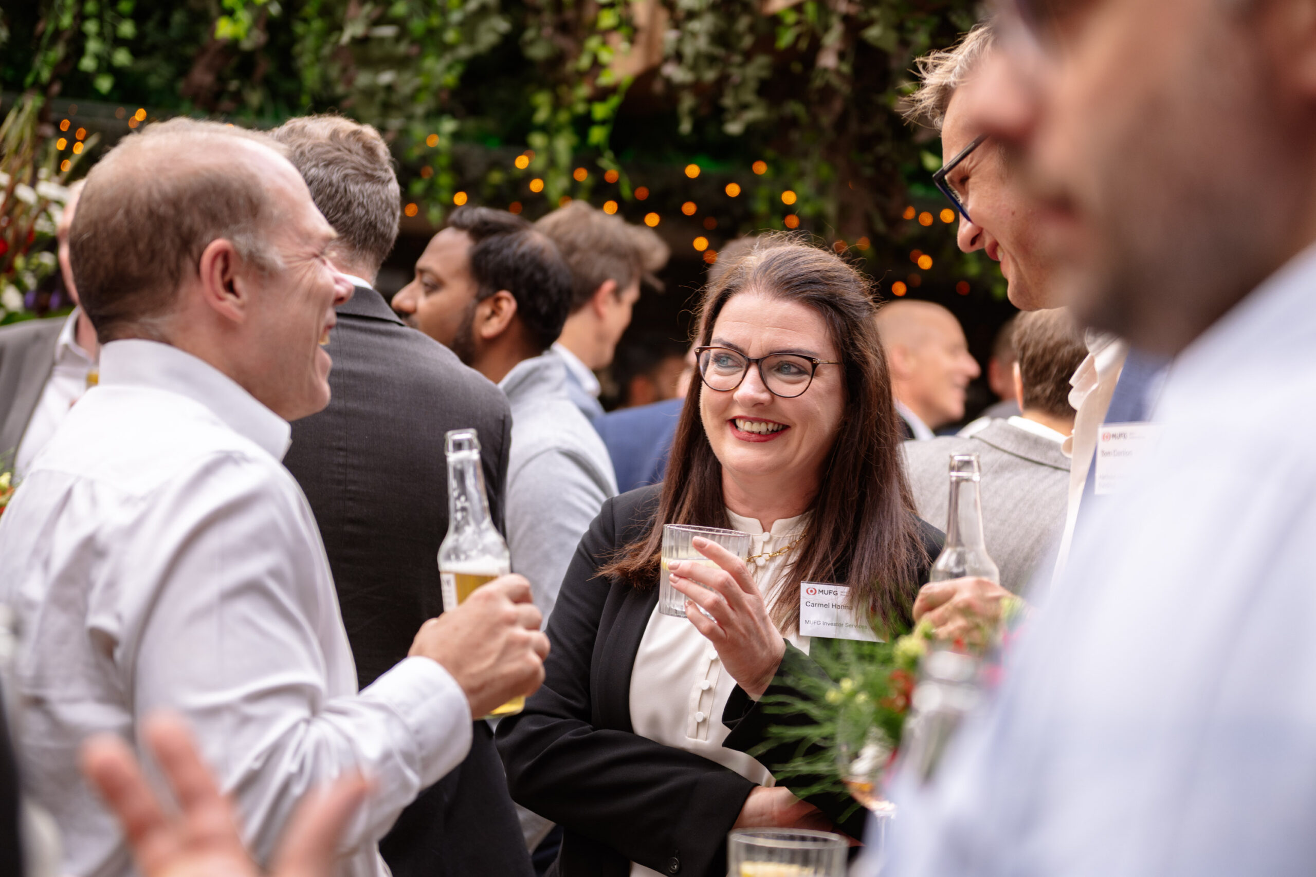 Group of coworkers at an outdoor reception, a smiling woman with glasses holds a drink at the center while others converse nearby.