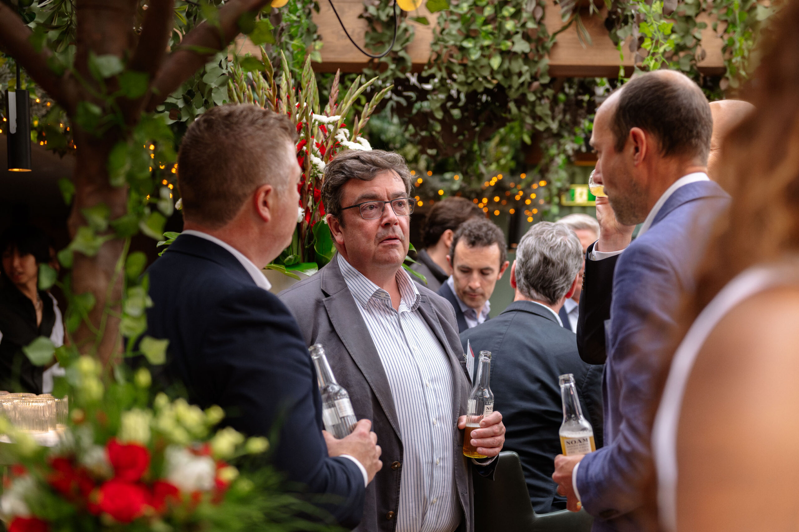 Group of guests in suits at a garden‑themed reception, holding beer bottles and chatting amid red floral arrangements.