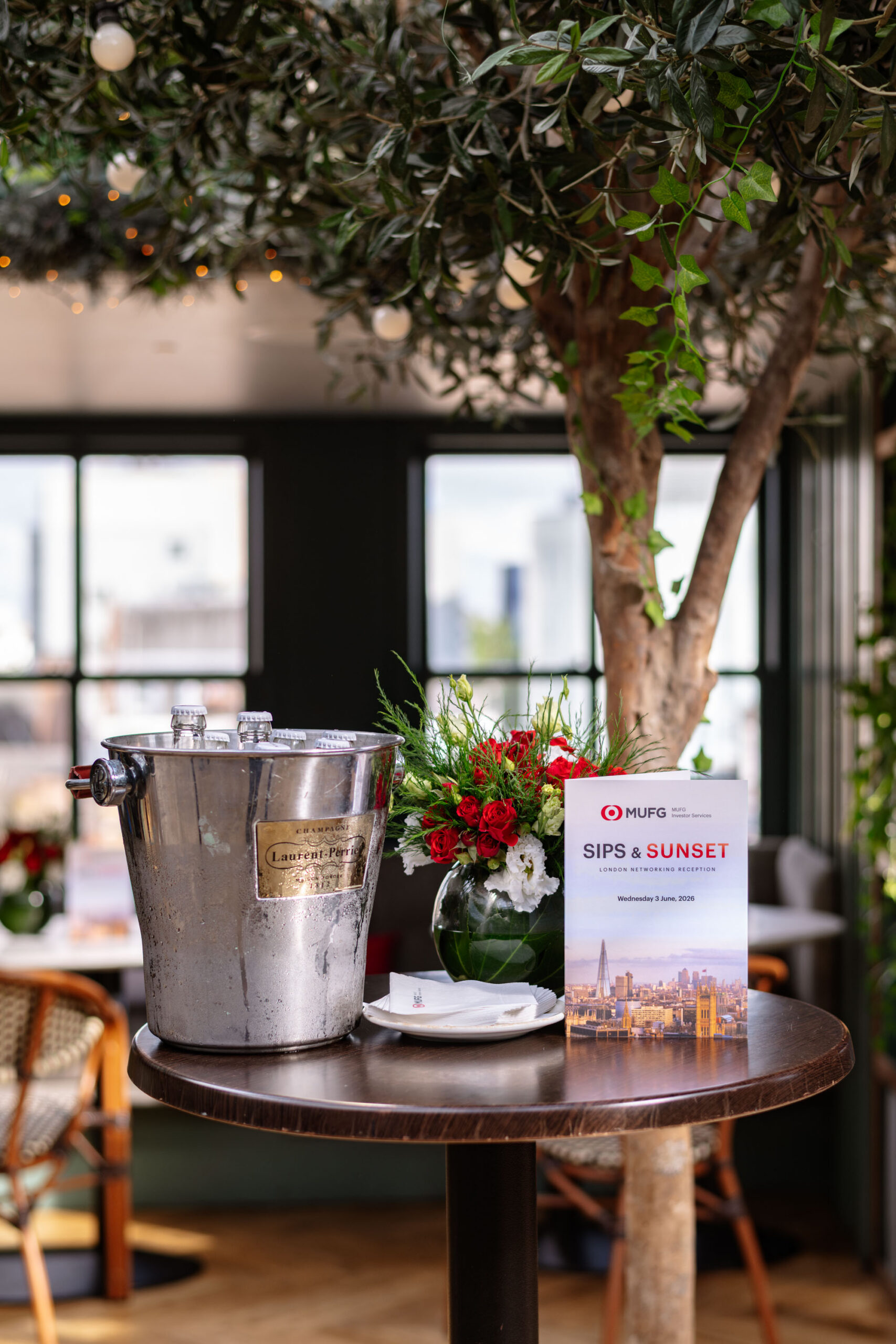Champagne cooler with ice on a round table beside a red-and-white flower bouquet and a 'Sips & Sunset' invitation in a bright indoor setting.