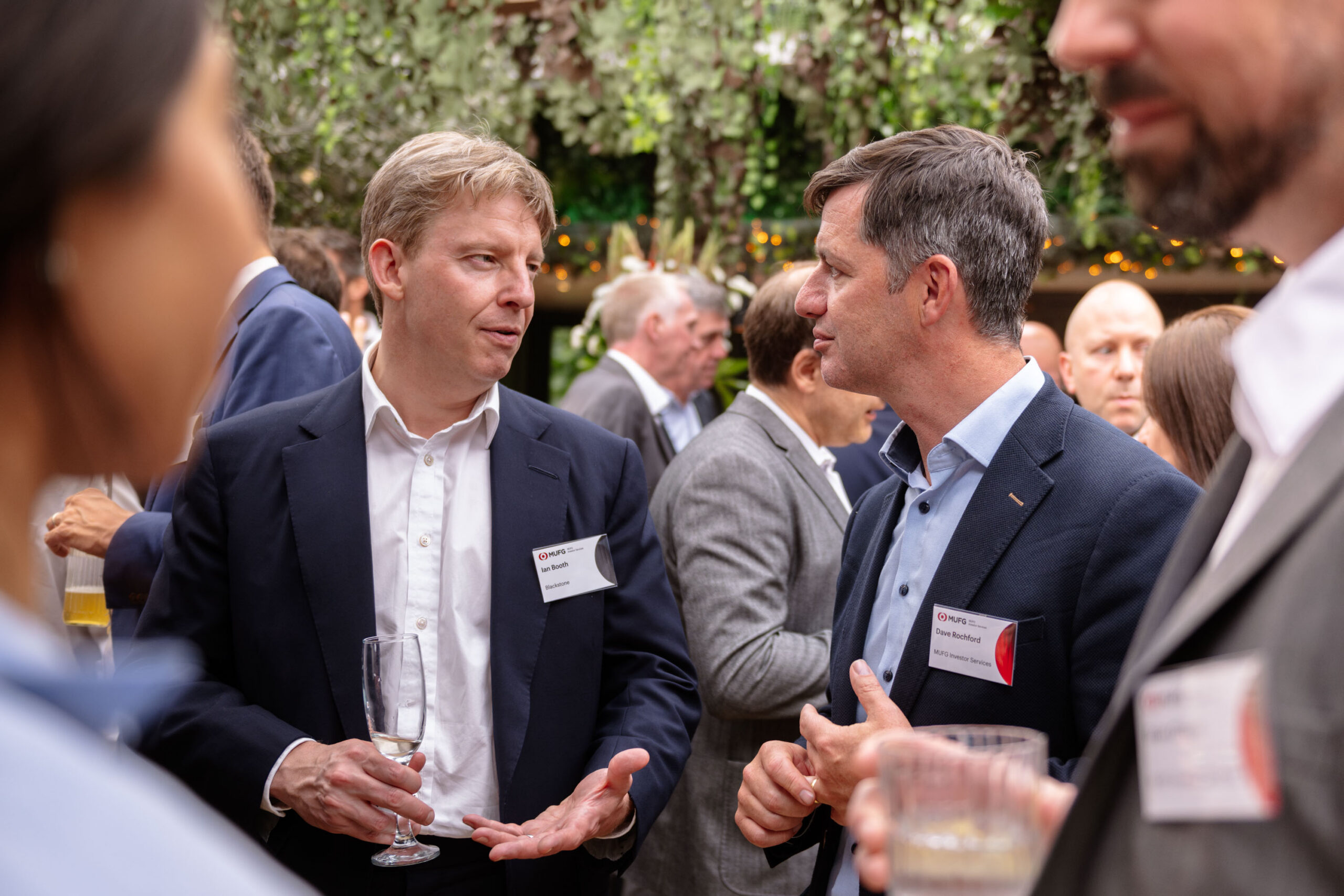 Two men in business suits discuss at a networking event, each holding a glass of drink with name badges visible on their jackets.