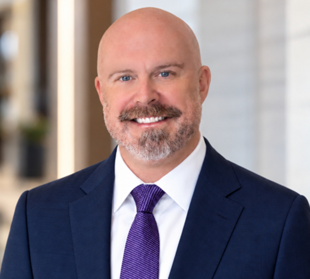 Professional headshot of a bald man with a trimmed beard in a navy suit, white shirt, and purple tie, smiling at the camera in a modern office setting.