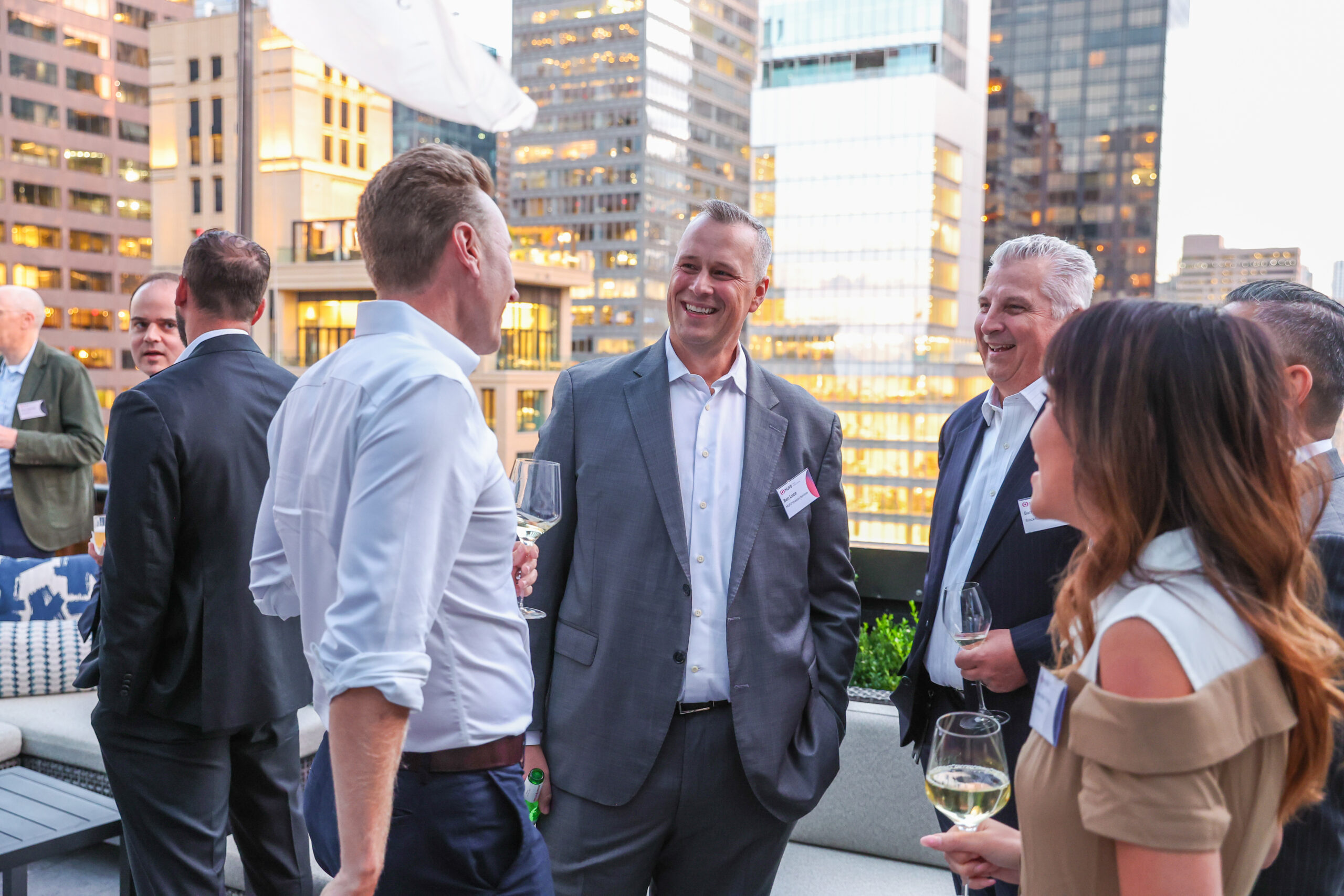 Group of professionals in suits chatting on a rooftop terrace in a city skyline setting, holding wine glasses at a networking event.