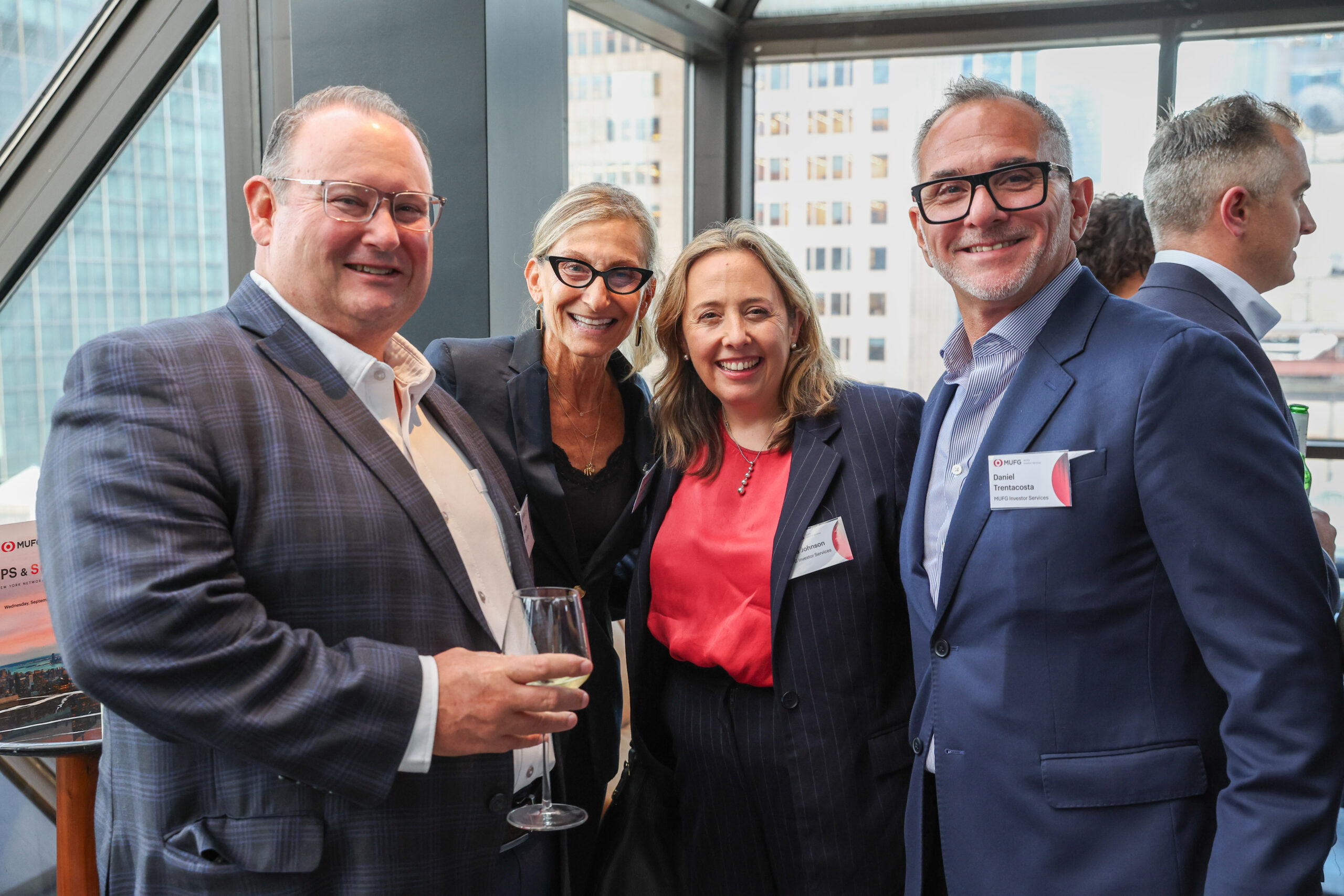 Group of four professionals in business attire posing and smiling at an event in a glass-walled venue, name tags visible on their jackets, one holding a wine glass