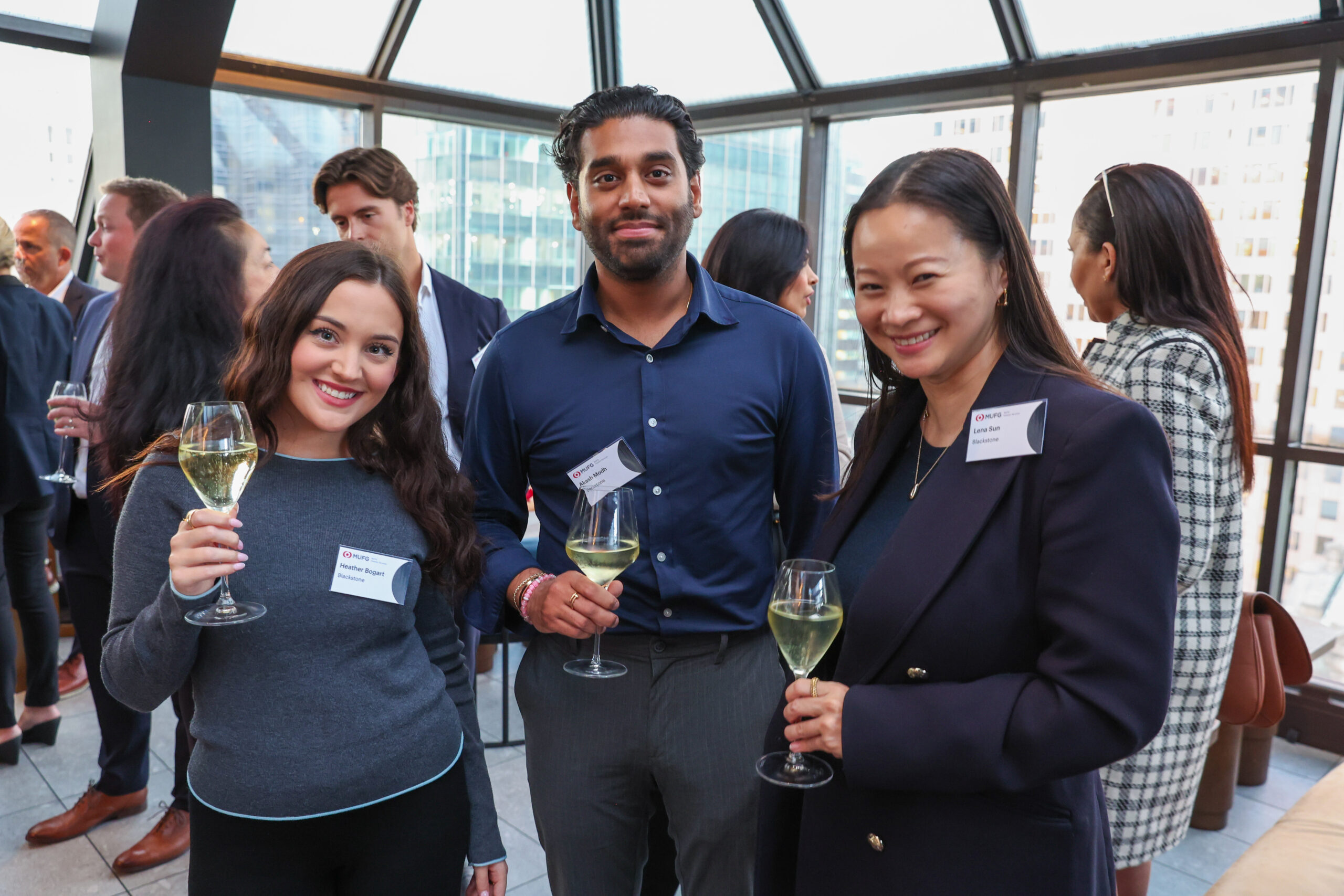 Group of professionals at a cocktail reception, smiling and holding wine glasses in a glass-walled venue.