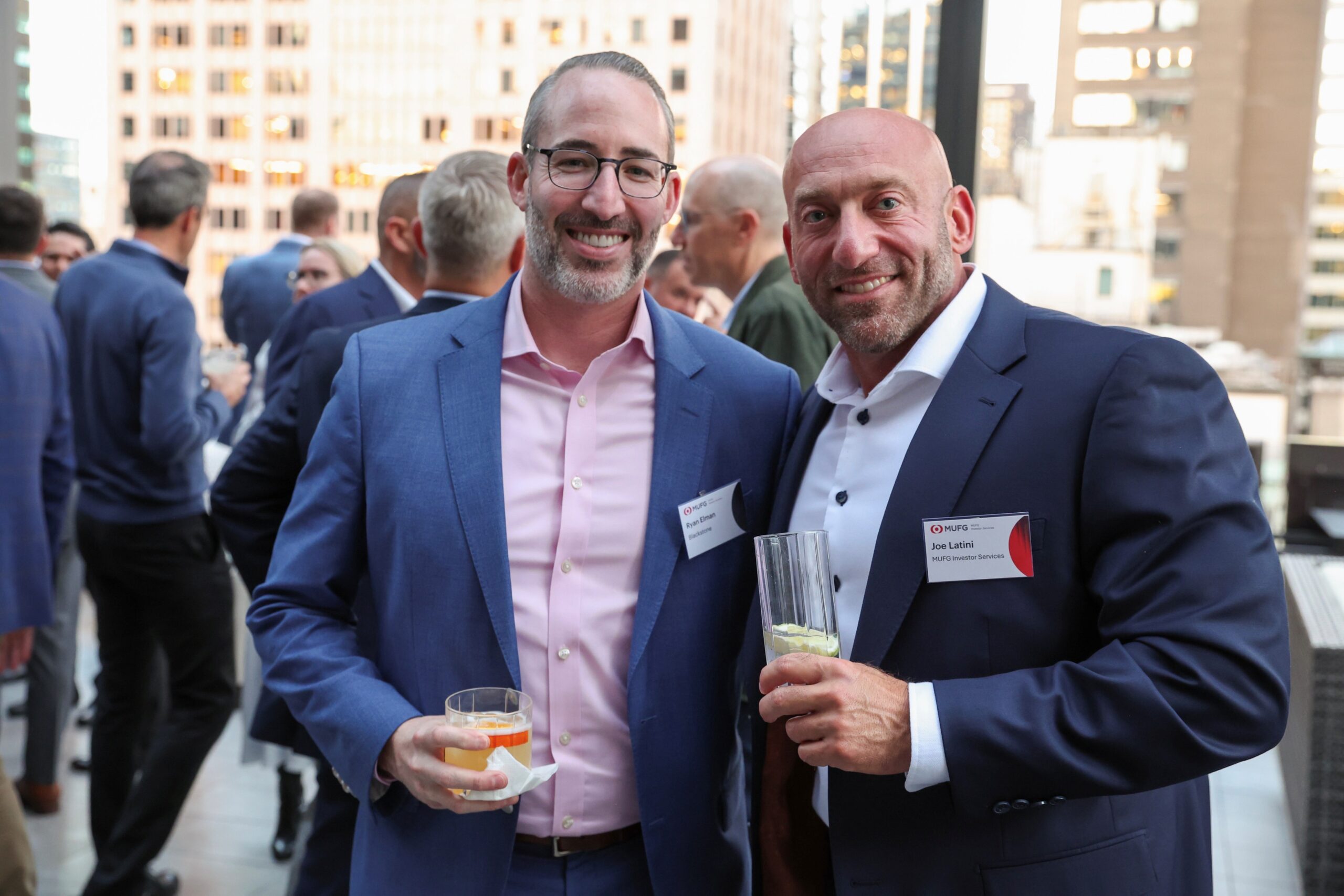 Two men in blue suits at a networking event, smiling and holding drinks with a cityscape behind them