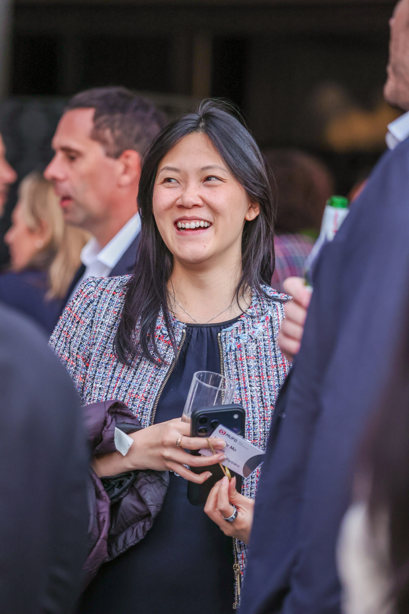 Smiling Asian woman at a social event, holding a wine glass and a smartphone with a name badge visible.
