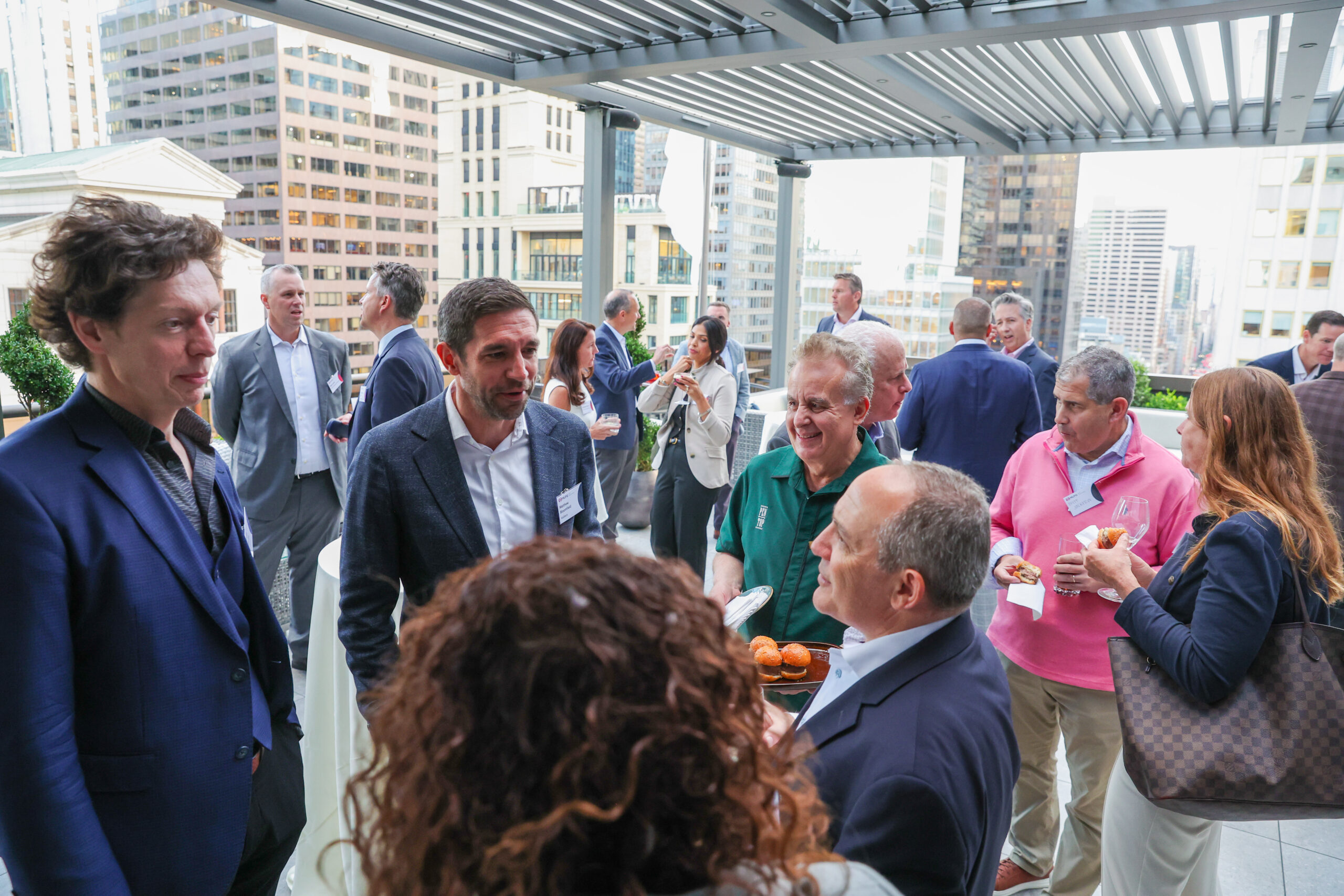 Group of professionals mingling on a city rooftop terrace, holding drinks and snacks with tall buildings in the background.