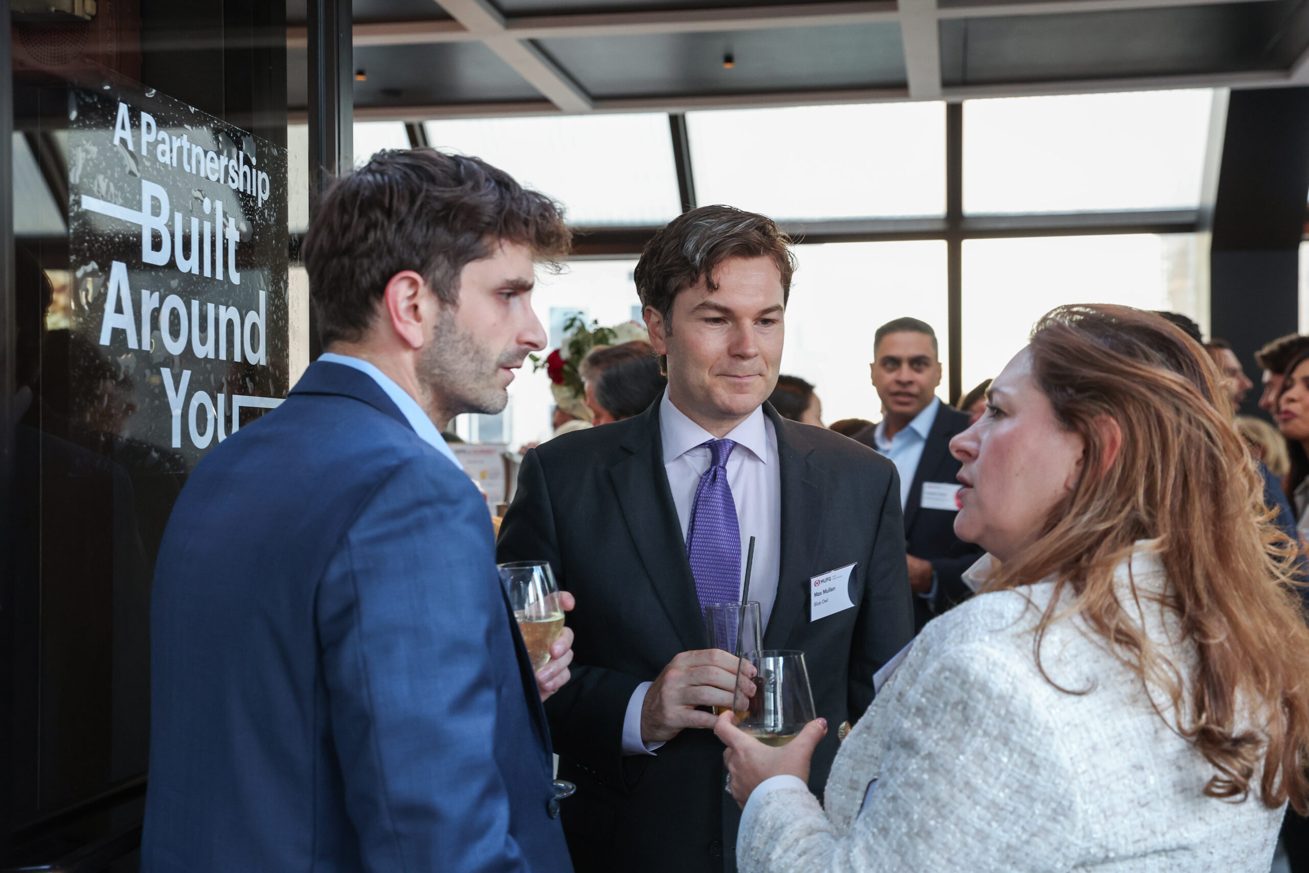 Group of professionals in suits and business attire converse at a networking event, holding glasses of champagne or wine in a modern venue.