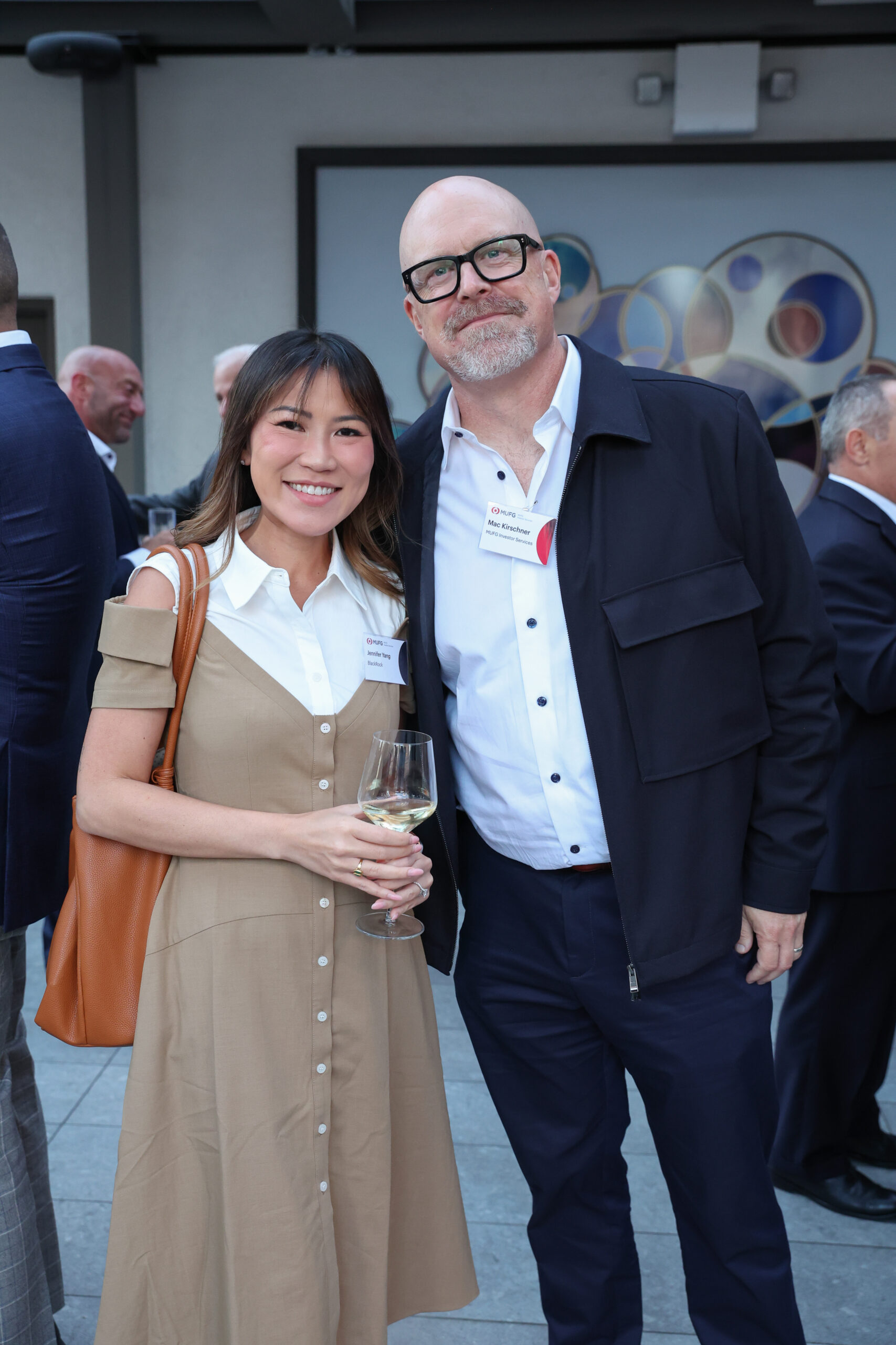 Two smiling adults posing together at an outdoor event; woman in a beige dress holding a wine glass, man in a dark jacket and white shirt wearing glasses and a name badge in front of a mural backdrop.