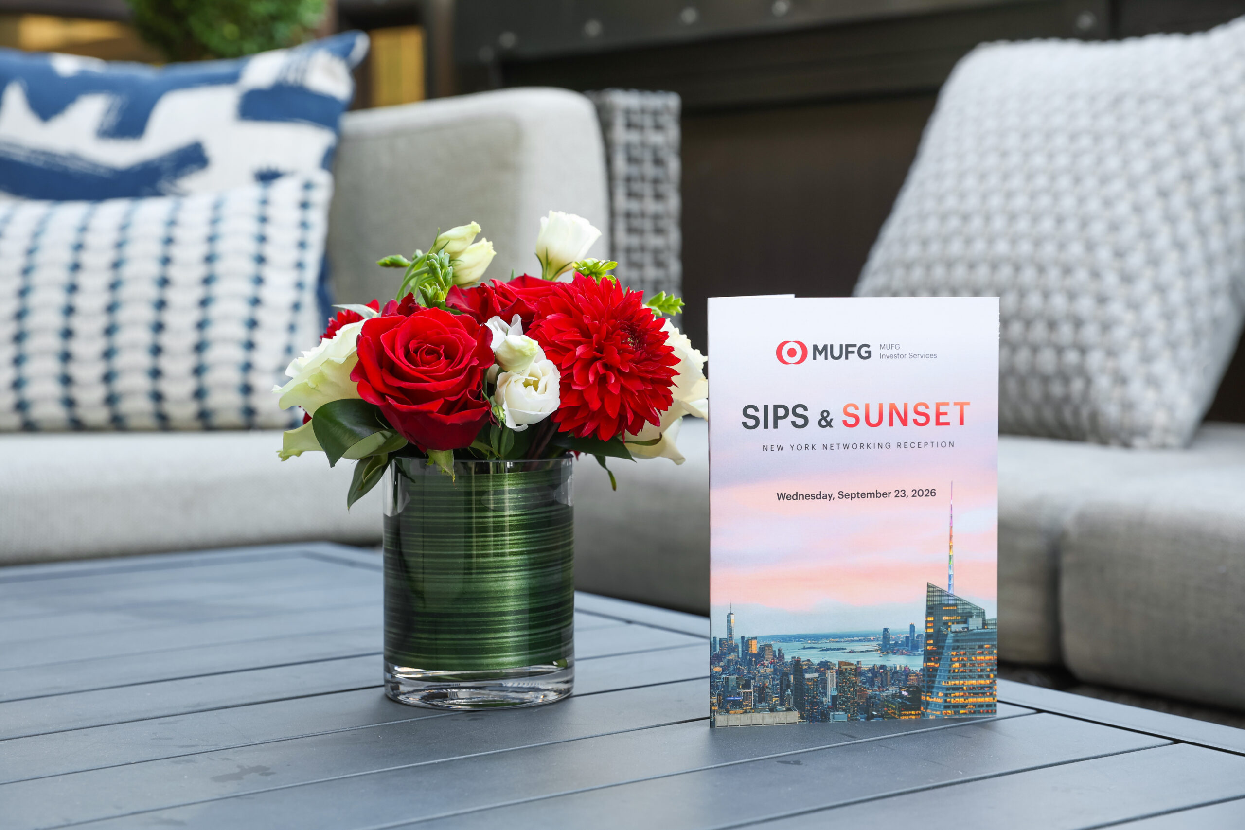 Bouquet of red roses and white flowers in a green glass vase on a gray outdoor table, with a MUFG SIPS & SUNSET invitation card nearby.