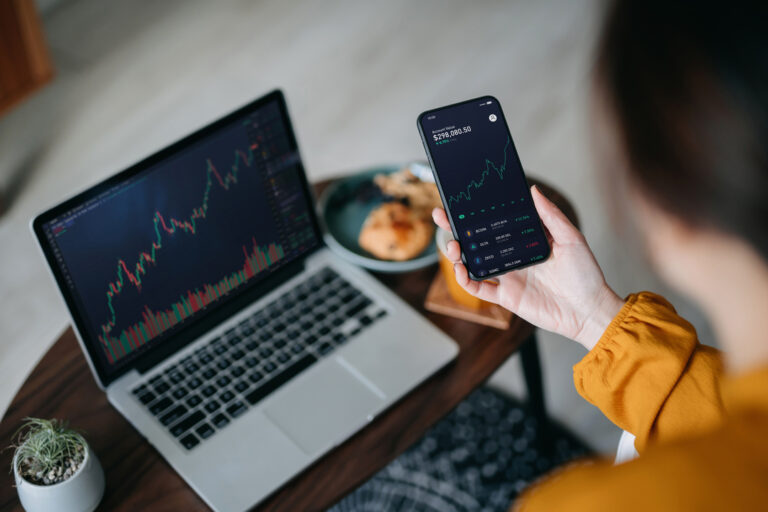 over the shoulder view of asian woman holding smartphone, analyzing investment trading data on crypto graph with smartphone and laptop while working at home. cryptocurrency, bitcoin, digital ledger