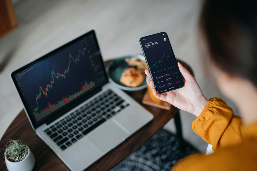 over the shoulder view of asian woman holding smartphone, analyzing investment trading data on crypto graph with smartphone and laptop while working at home. cryptocurrency, bitcoin, digital ledger