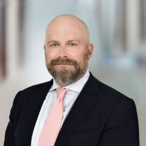 Professional headshot of a bald man with a beard, in a navy suit and pink tie, blurred office backdrop.
